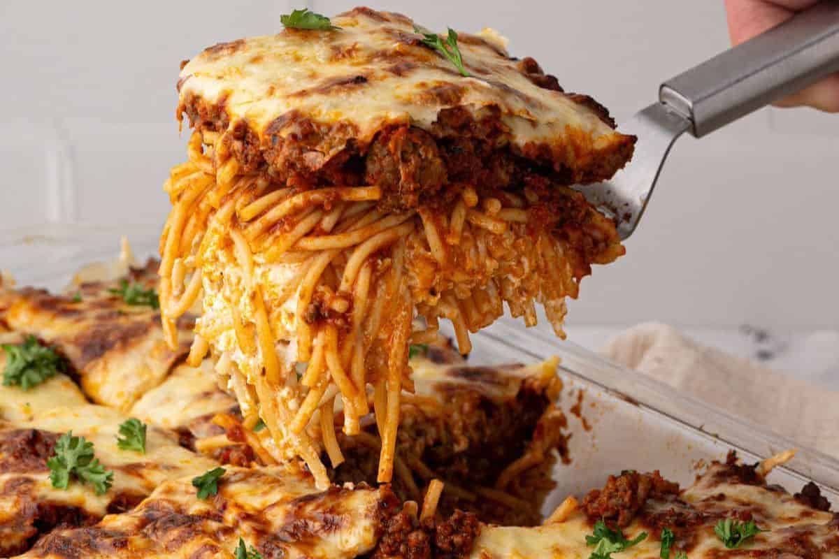 A spatula lifting a cheesy, layered slice of Million Dollar Spaghetti from the casserole dish