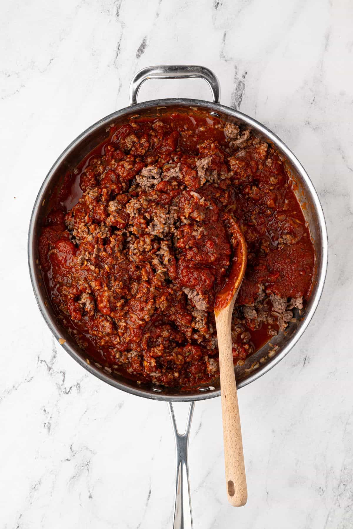 A skillet filled with cooked ground beef mixed with marinara sauce, with a wooden spoon stirring it.