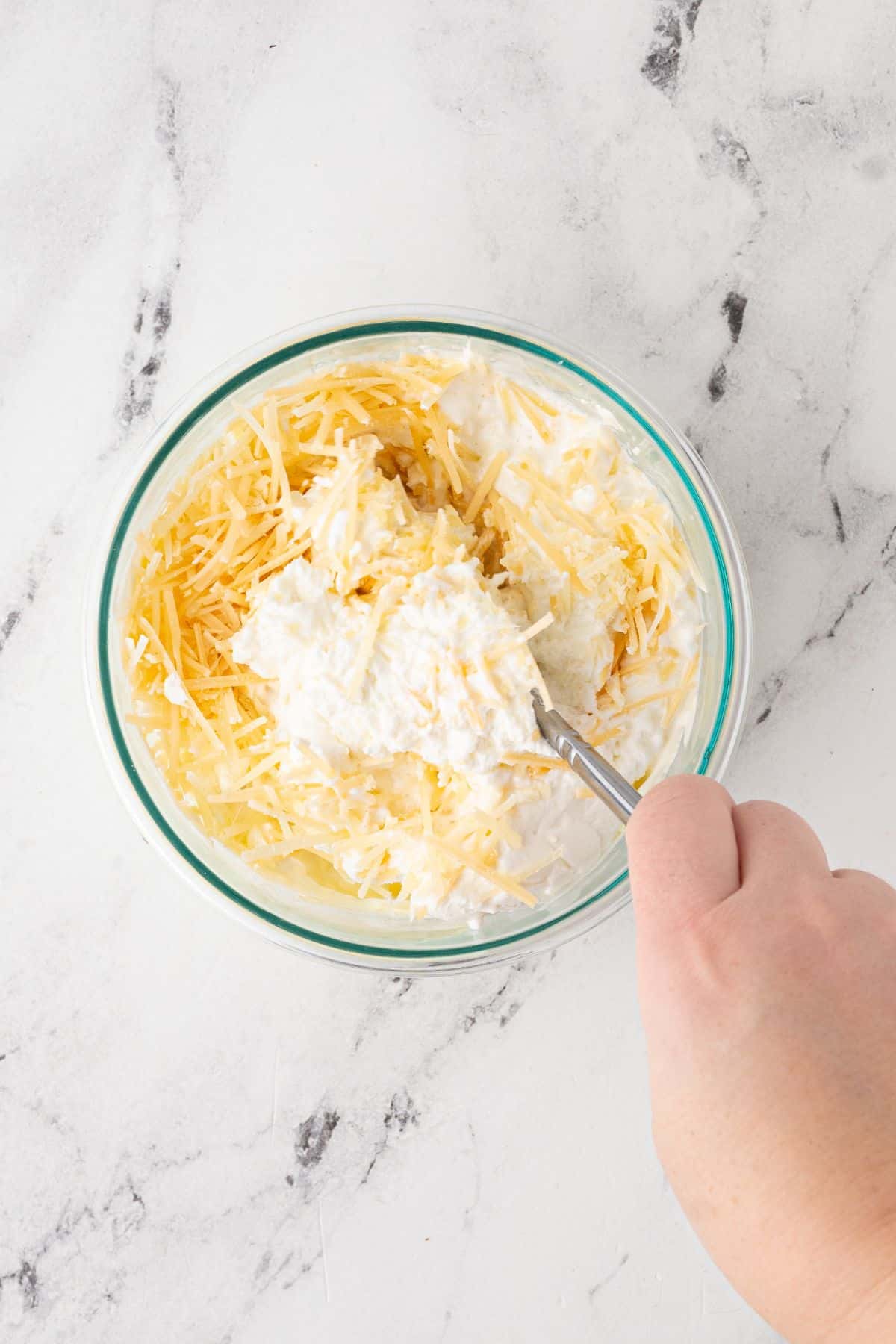 A hand stirring a bowl of ricotta, cottage cheese, and shredded cheese.