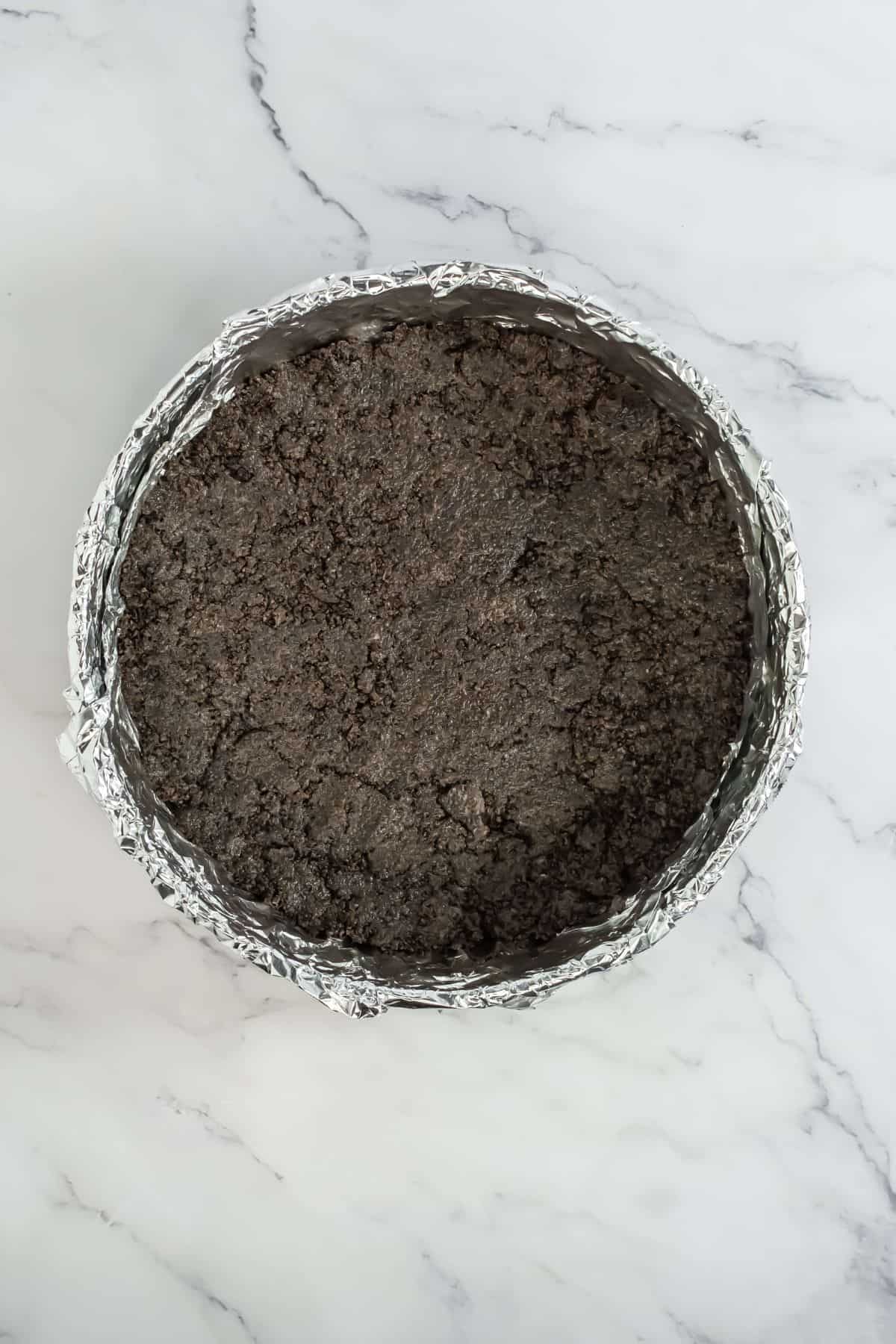 Crumbled Oreo crust pressed into a springform pan, lined with foil for easy removal.