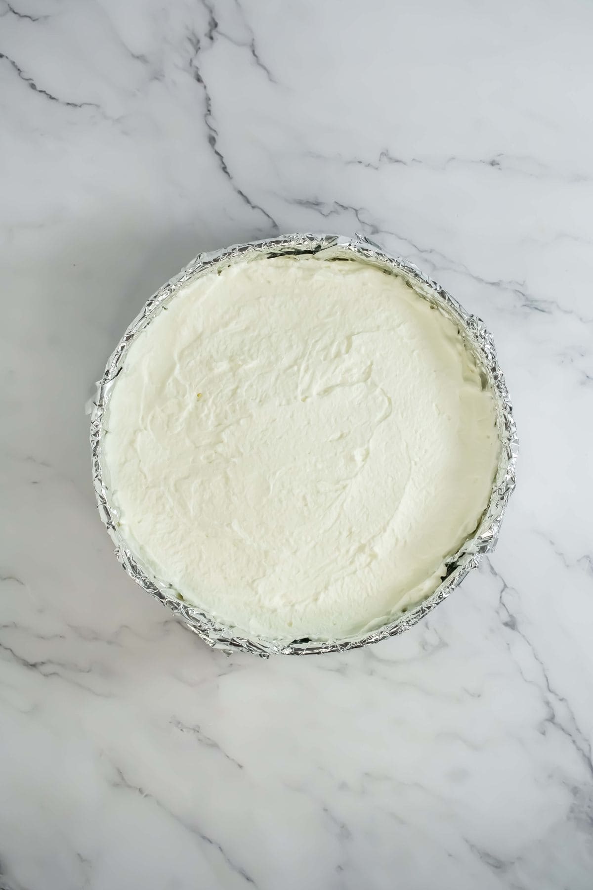 A springform pan lined with foil, containing a smooth white whipped cream layer spread evenly.