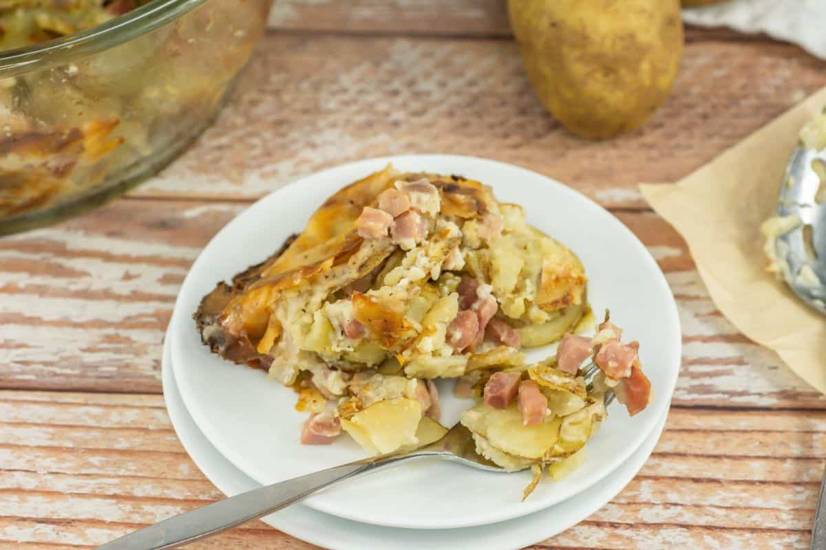 Serving of scalloped potatoes and ham on a white plate with a fork, with raw potatoes and a bowl with more scalloped potatoes in the background