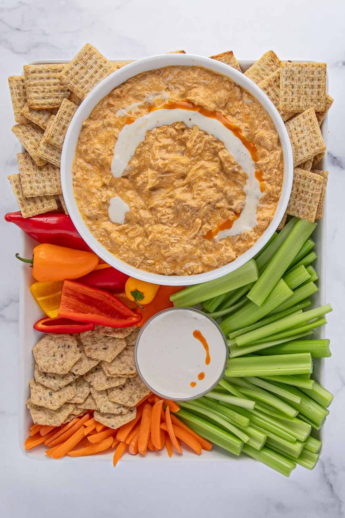 a white rectangular tray filled with snacks, a small bowl of ranch dressing, and a large bowl of slow cooker buffalo chicken dip