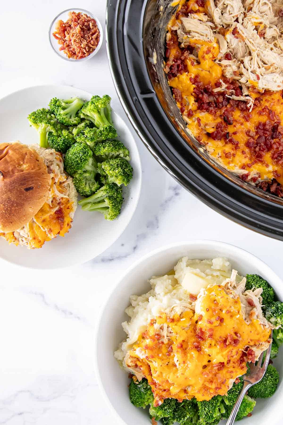 Overhead shot of slow cooker with slow cooker crack chicken, a bowl of mashed potatoes topped with crack chicken paired with a side of broccoli, and a crack chicken sandwich with a side of broccoli on a plate