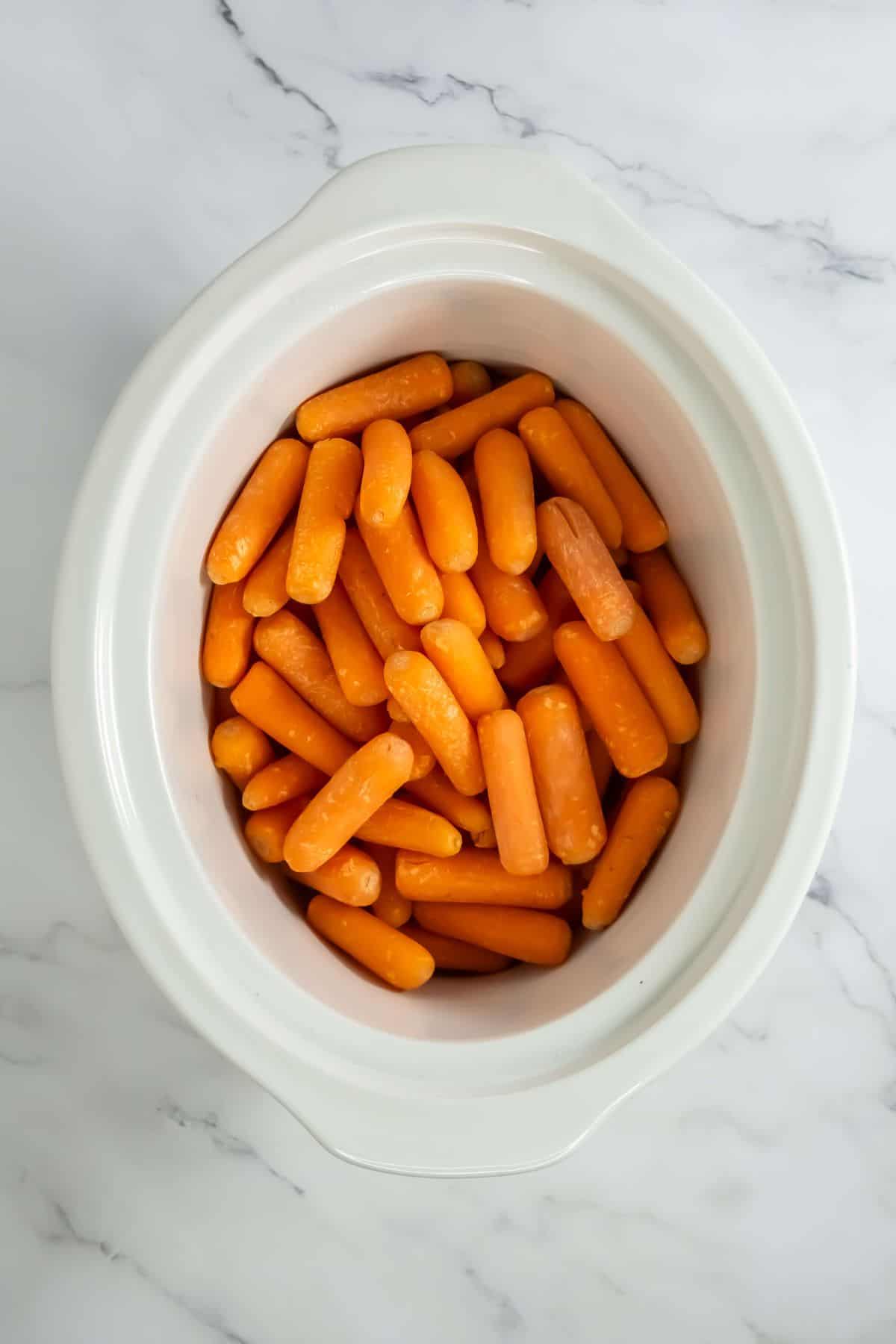 A slow cooker filled with fresh, uncooked baby carrots, ready for the glaze.