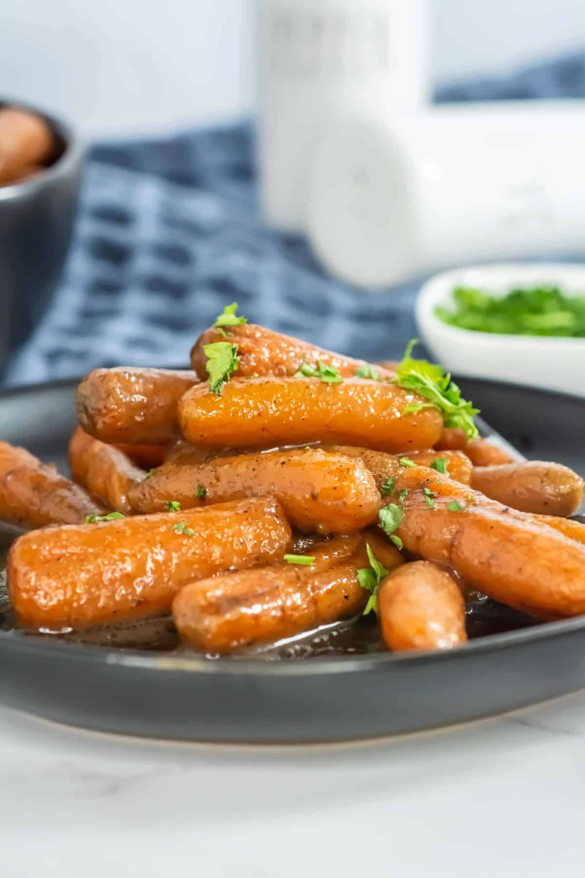 Close-up side view of glossy glazed carrots, garnished with parsley, served on a black plate. A small dish of parsley and blue napkin are visible in the background.
