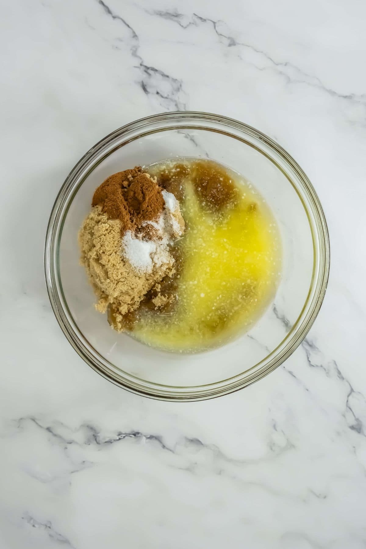 A glass mixing bowl containing ingredients for the glaze, including melted butter, brown sugar, and spices. The ingredients are unmixed and visually distinct.