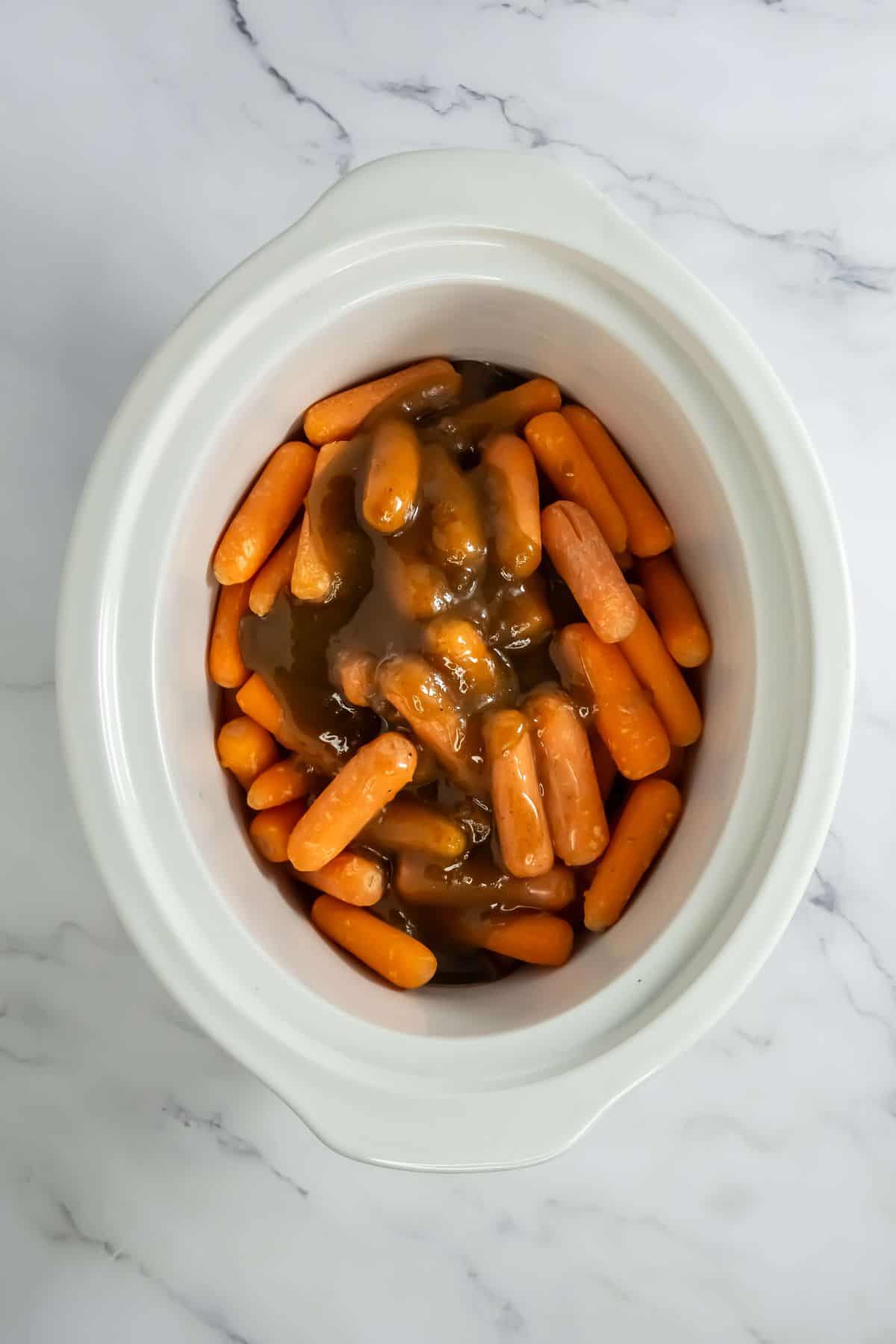 A slow cooker filled with uncooked baby carrots, just after the brown glaze has been poured over them. The glaze is thick and glossy, coating the carrots before cooking begins.