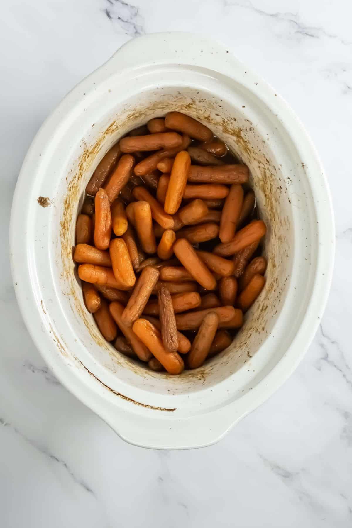 A slow cooker filled with cooked baby carrots coated in a brown glaze. The carrots look tender, and the sides of the slow cooker show light caramelized glaze residue.