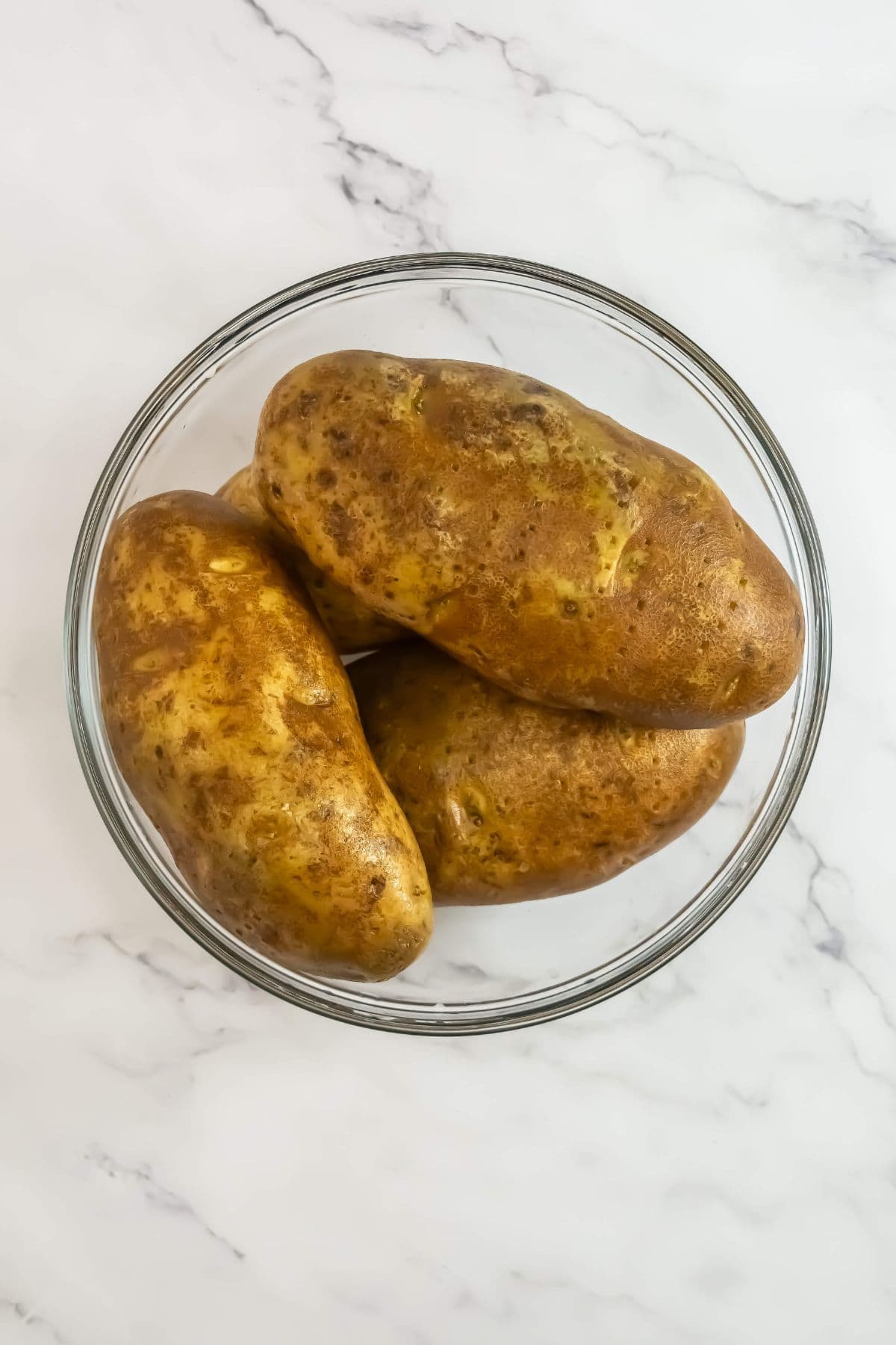 Three raw russet potatoes are placed in a clear glass bowl. The potatoes are unpeeled, showcasing their textured, brown skin. The background is a clean, white marble surface