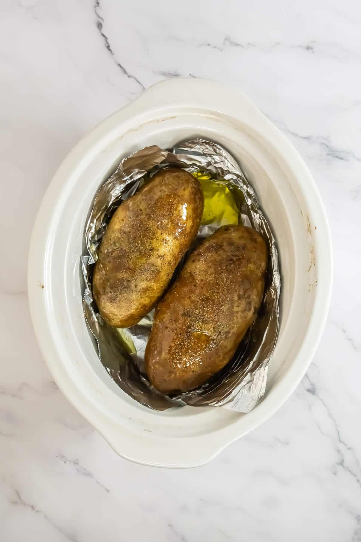 A closer top-down view shows two jacket potatoes in the slow cooker. The potatoes appear glistening, likely due to oil, and their skins are more browned and crispy.