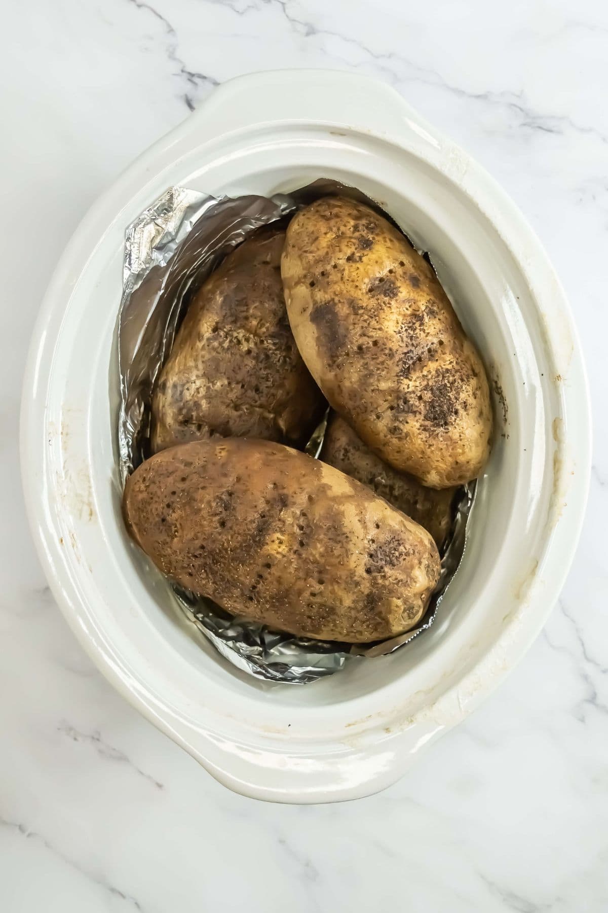 A top-down view of the slow cooker shows three seasoned potatoes. The aluminum foil lining is visible, and the potatoes have started to brown and crisp, indicating they are partway through the cooking process.