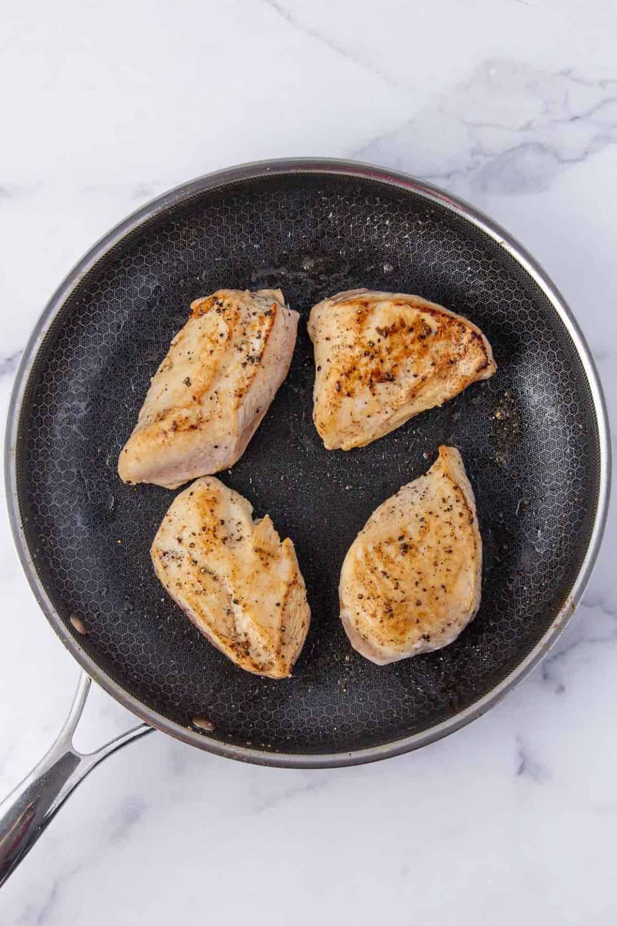 Four seasoned chicken breasts are searing in a nonstick skillet, showing a light golden-brown color and grill marks.