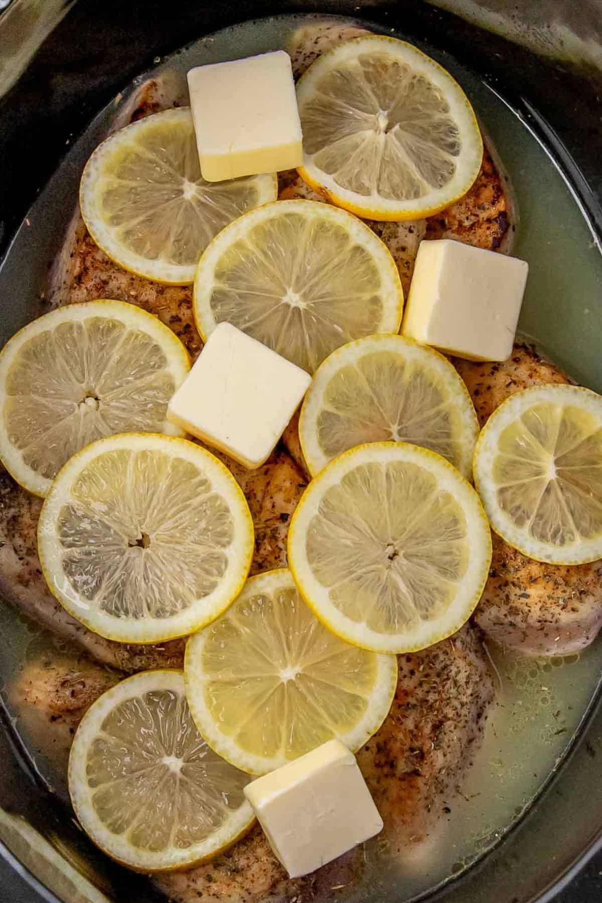 A close-up of seasoned chicken breasts in the slow cooker topped with fresh lemon slices and butter cubes, ready for cooking.