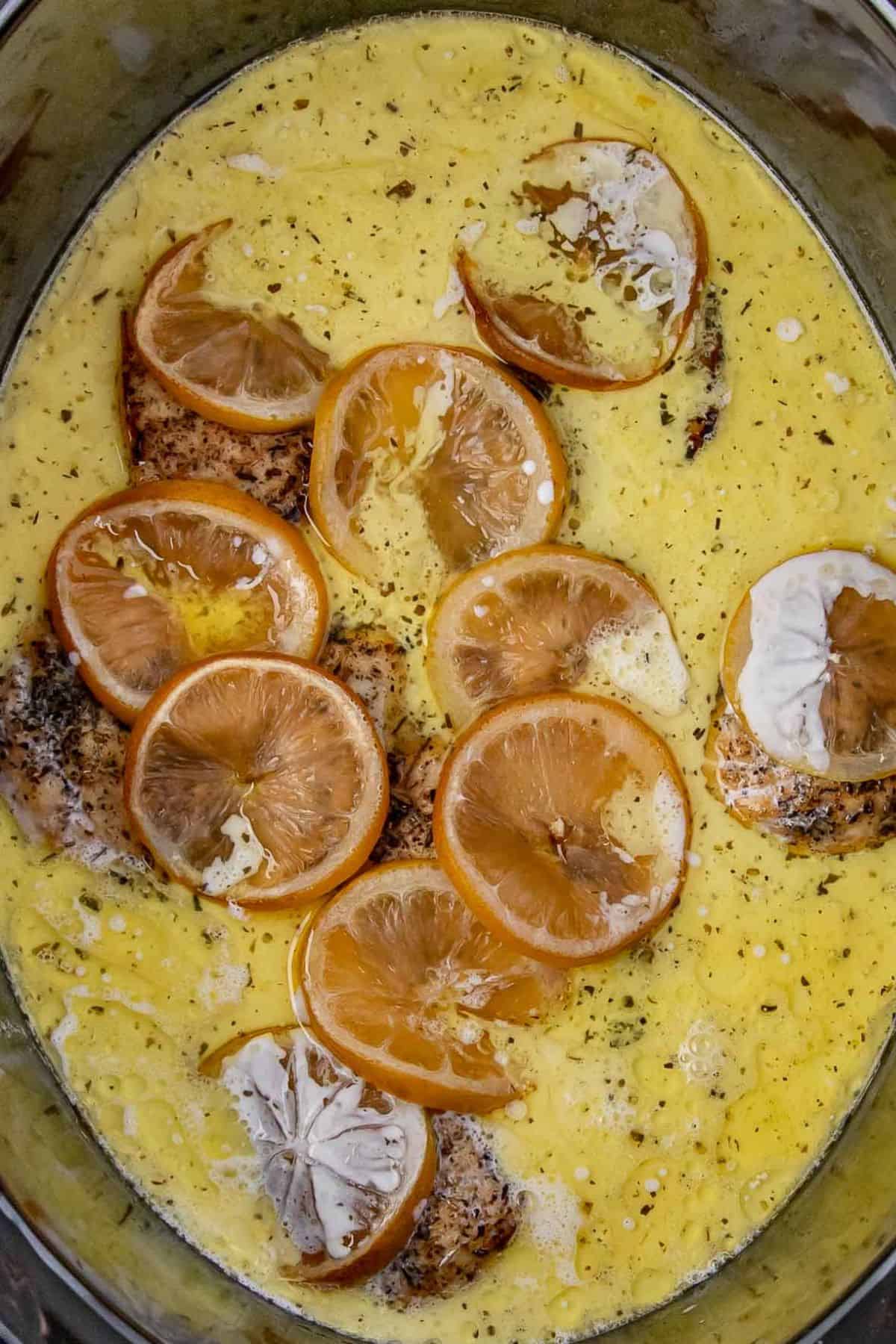 A close-up of chicken breasts in a slow cooker, topped with lemon slices and swimming in a vibrant yellow, creamy lemon sauce with herbs.