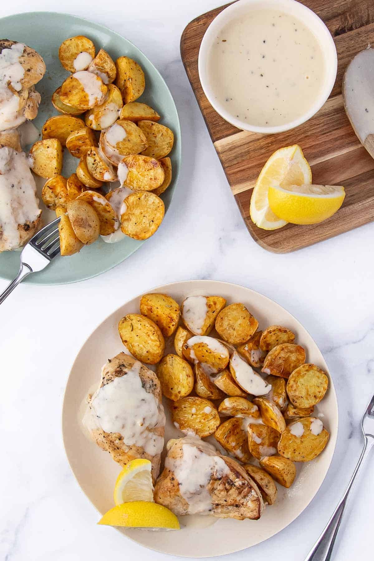 A top-down view of two plates with lemon chicken, roasted potatoes drizzled in creamy sauce, a fork in one plate, and a wooden cutting board holding lemon wedges and a sauce ramekin.