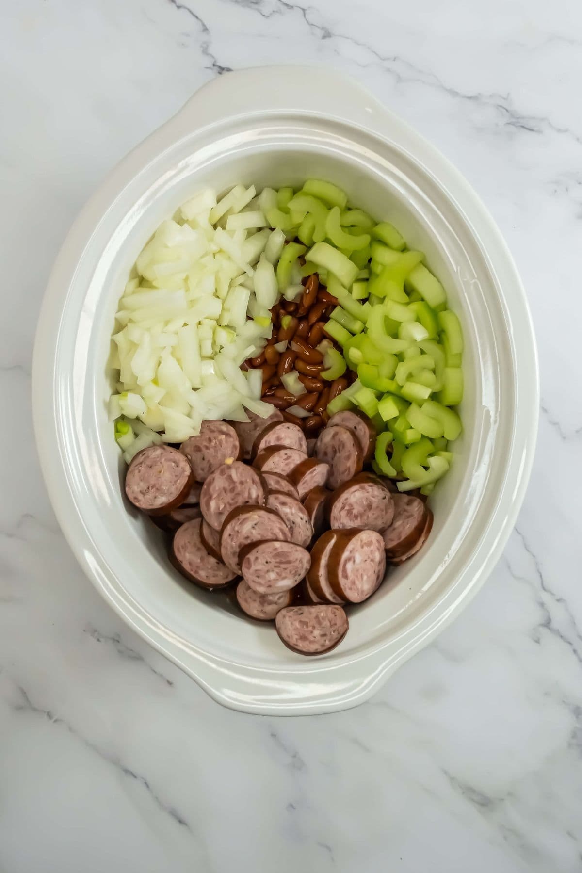 Sliced sausage, beans, celery, and onion layered in a slow cooker.