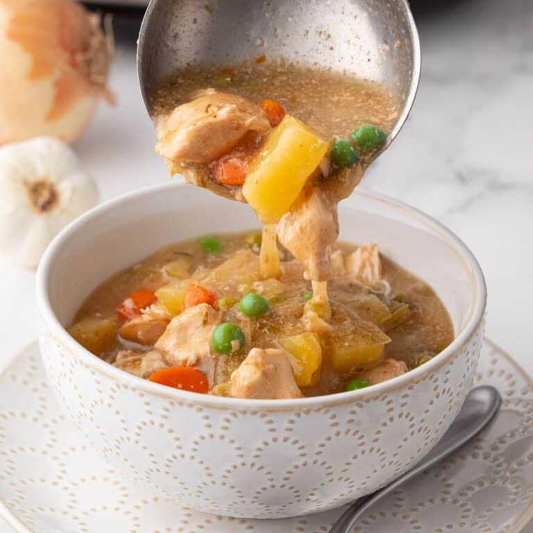 A bowl of crock pot chicken stew with vegetables like carrots, peas, and potatoes, being ladled into a decorative white bowl.