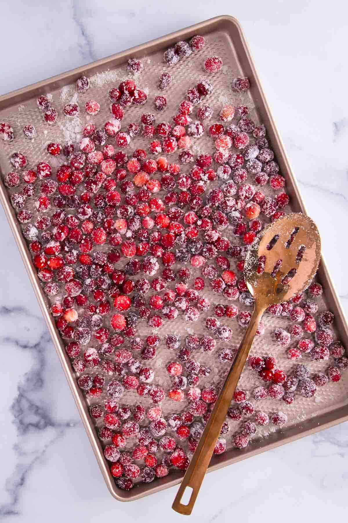 a sheet pan filled with cranberries coated in sugar, and a slotted copper spoon rests on the sheet