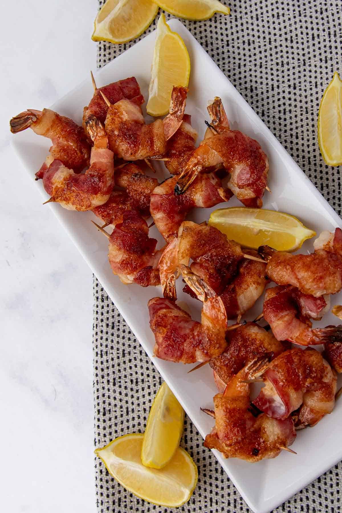 crispy Bacon wrapped shrimp arranged neatly on a white serving plate with lemon wedges, set against a patterned fabric background.