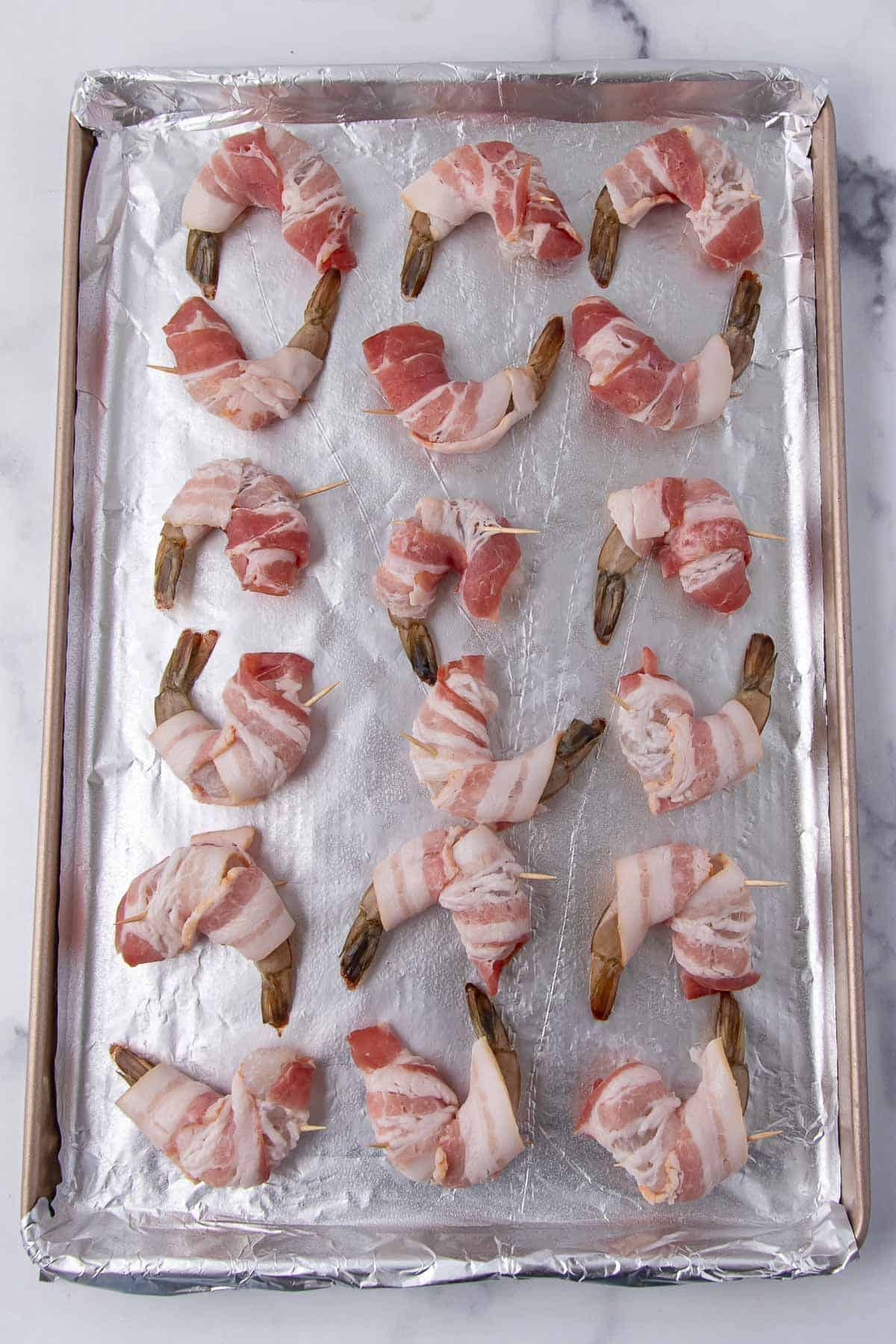 Shrimp wrapped in bacon and secured with toothpicks, arranged on a foil-lined baking tray.