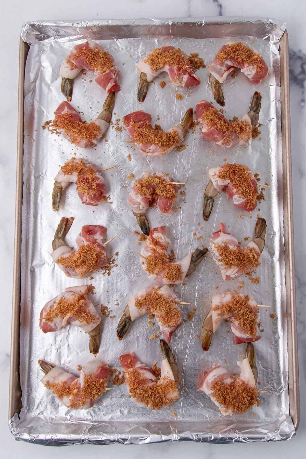Bacon-wrapped shrimp topped with a brown sugar mixture on a foil-lined baking tray, prepared for baking.