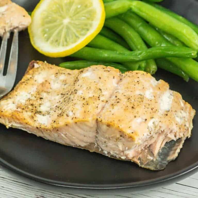 A single serving of broiled salmon with green beans and a lemon wedge on a black plate.