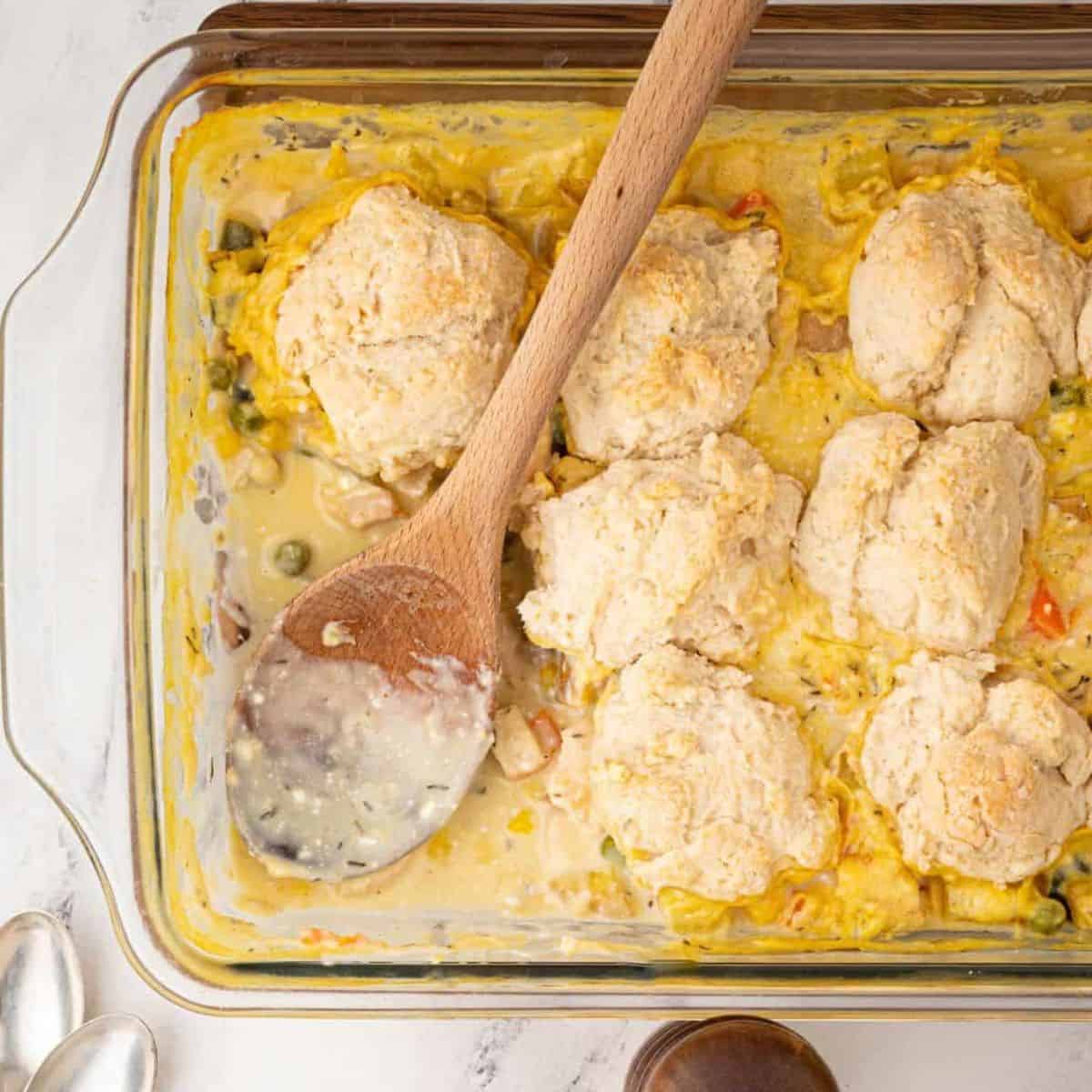 chicken pot pie casserole in a 9x13 casserole dish with a biscuit topping and a wooden spoon.