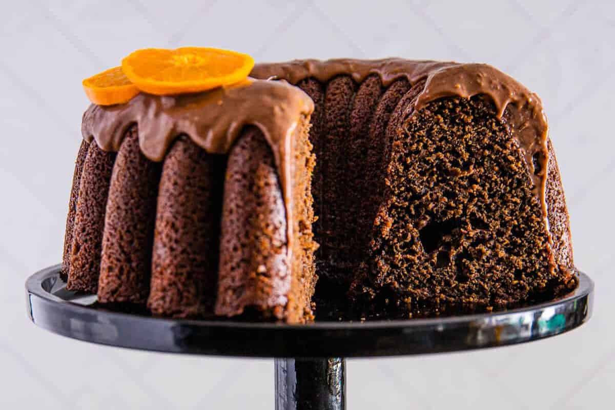 chocolate orange cake with chocolate ganache and orange slices on top, with a slice removed to reveal the moist interior, all on a black cake stand