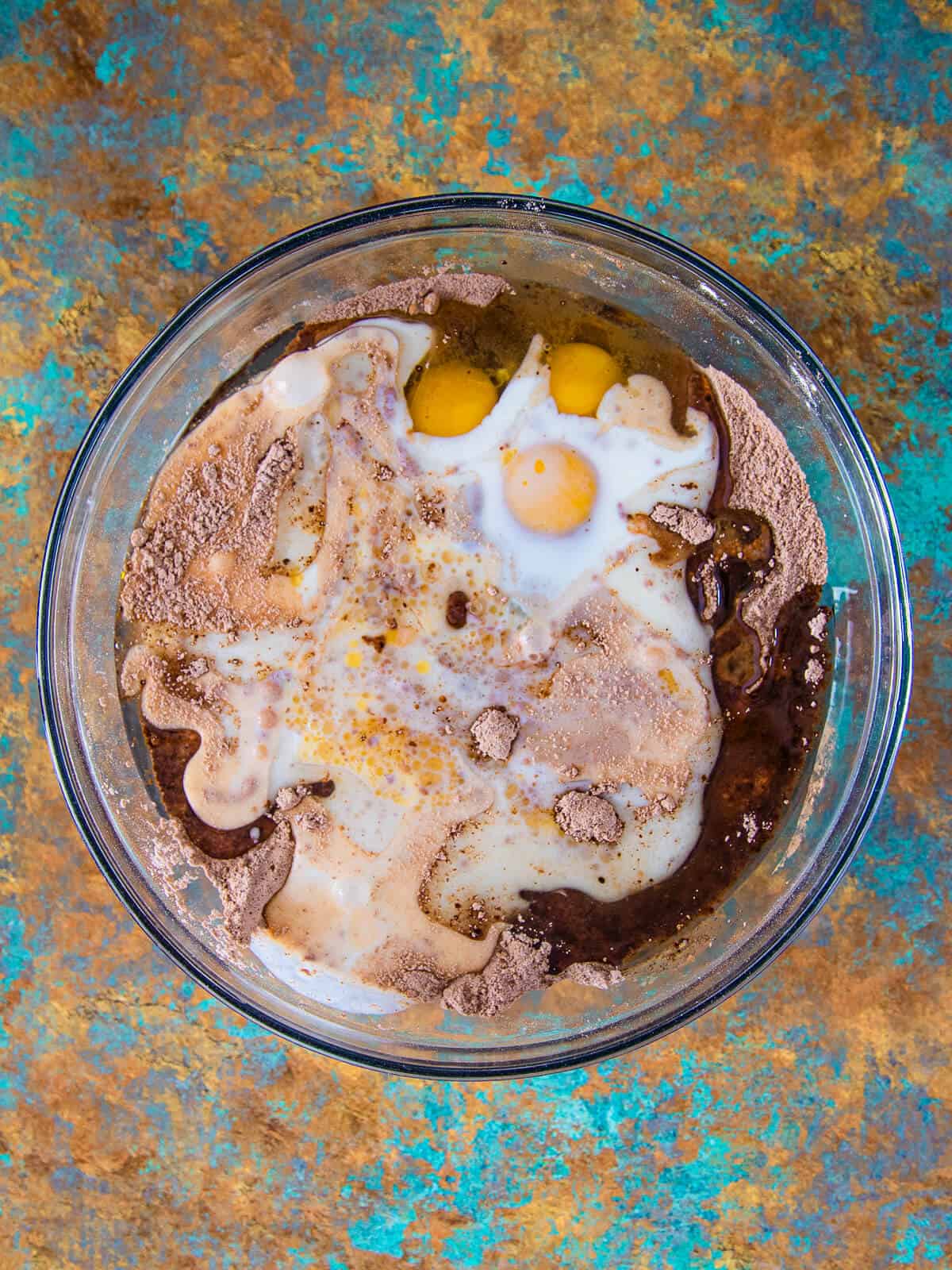 A top-down view of a mixing bowl containing cocoa powder, flour, eggs, milk, and other wet ingredients before mixing.