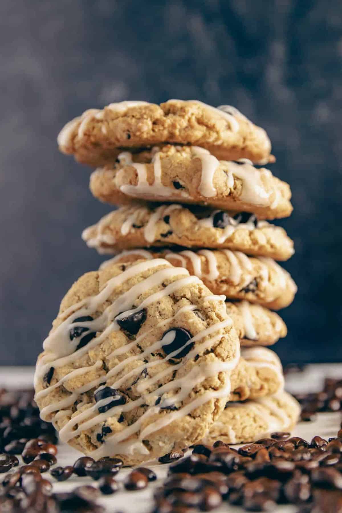 A stack of coffee cookies with white icing drizzle, surrounded by scattered coffee beans, against a dark background