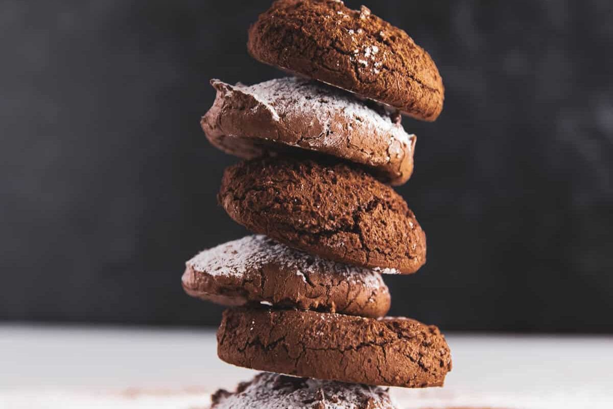 A tall stack of five fudgy chocolate cookies, some dusted with powdered sugar, against a dark background