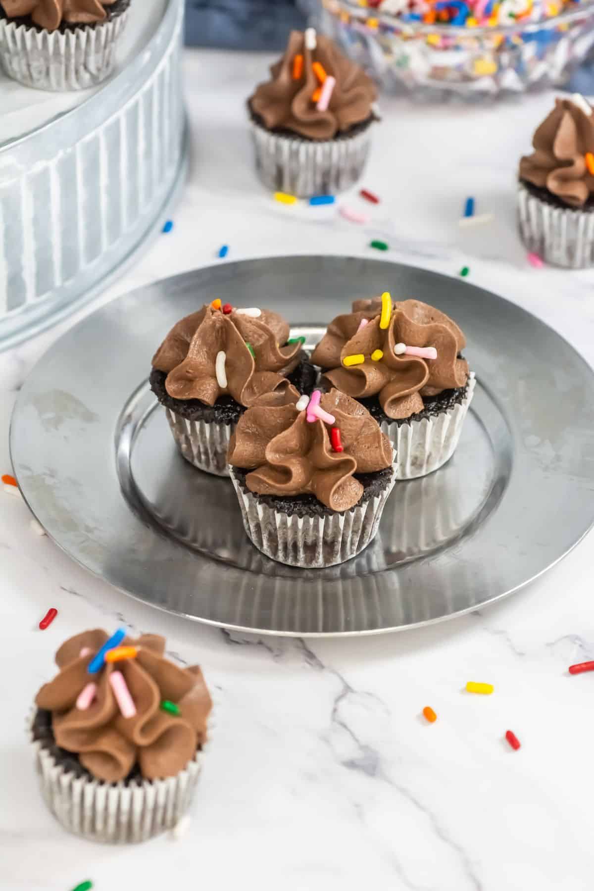 Three mini chocolate cupcakes with chocolate frosting and sprinkles on a silver plate, with scattered sprinkles around them