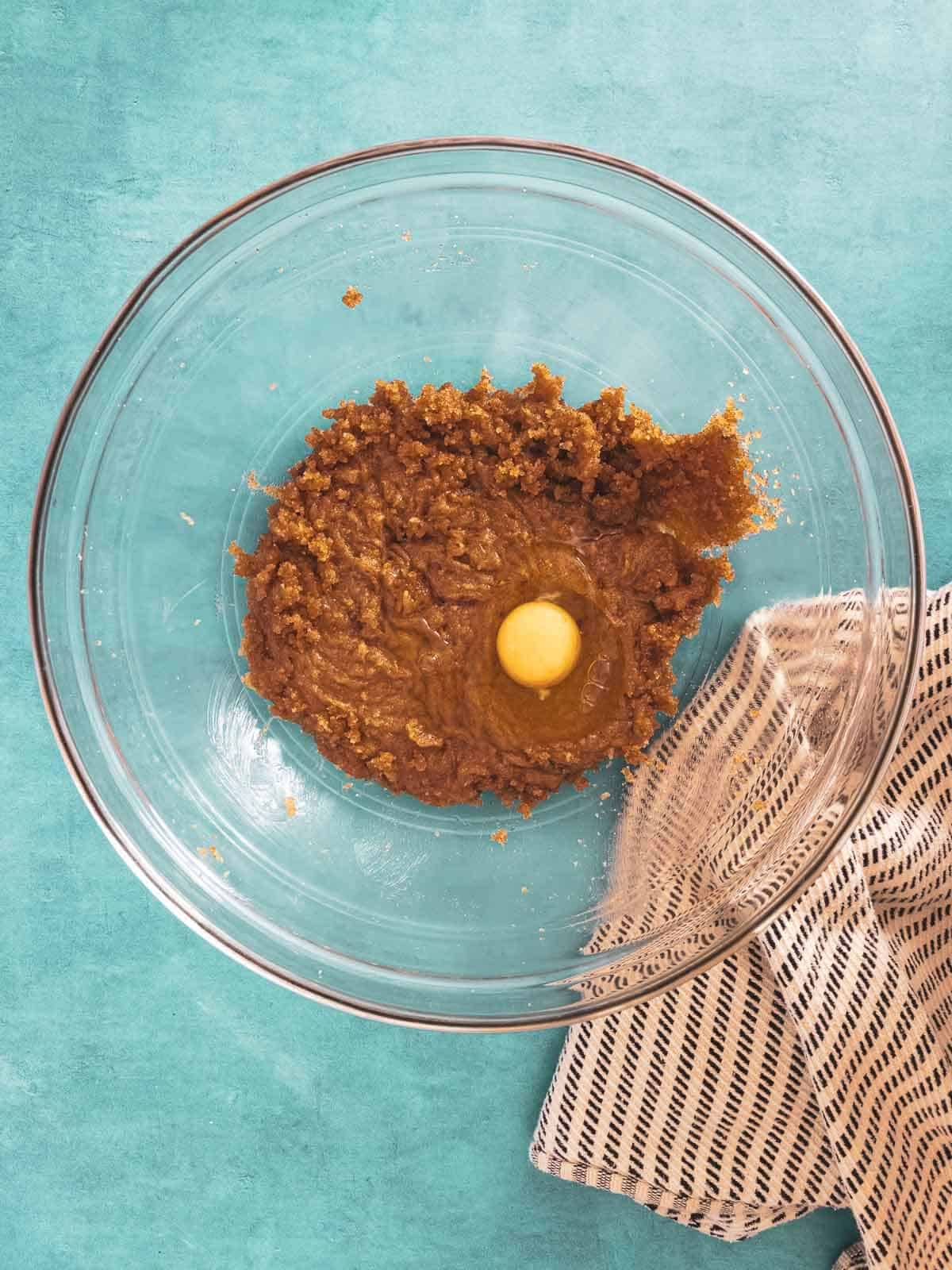 A mixing bowl with brown sugar and an egg being combined on a vibrant turquoise background.