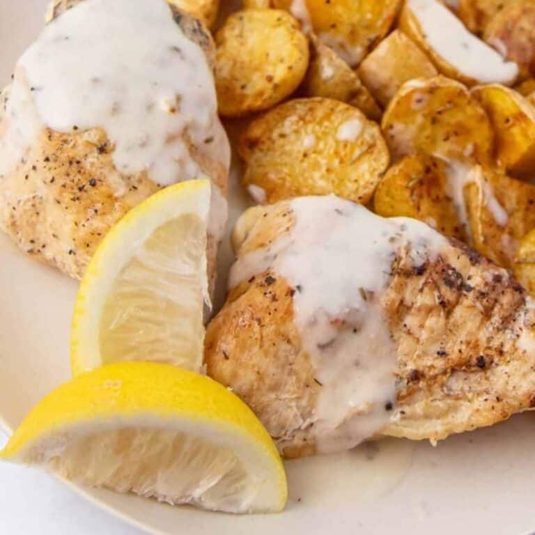 two slow cooker lemon chicken breasts on a plate with a cream sauce, lemon wedges for garnish and a side of roasted potatoes