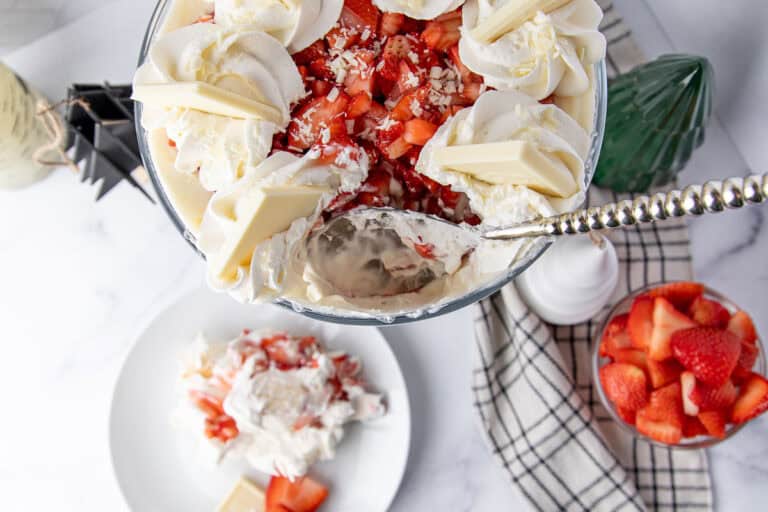 Strawberry trifle with white chocolate with a serving spoon dipped into it, surrounded by a plaid kitchen towel, a bowl of chopped strawberries, a decorative green glass tree, and a white plate with a served portion of the trifle