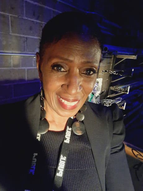 Smiling professional woman in a dark blazer at an event with blue lighting and equipment in the background.