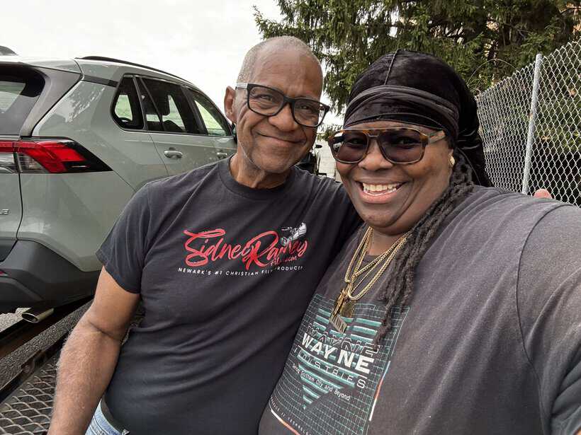 Smiling man and woman taking a selfie outdoors with a car in the background.