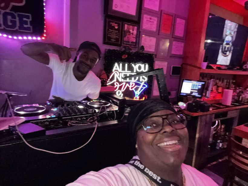 DJ performing at a lively nightclub with vibrant neon lights and a happy guest taking a selfie in front.