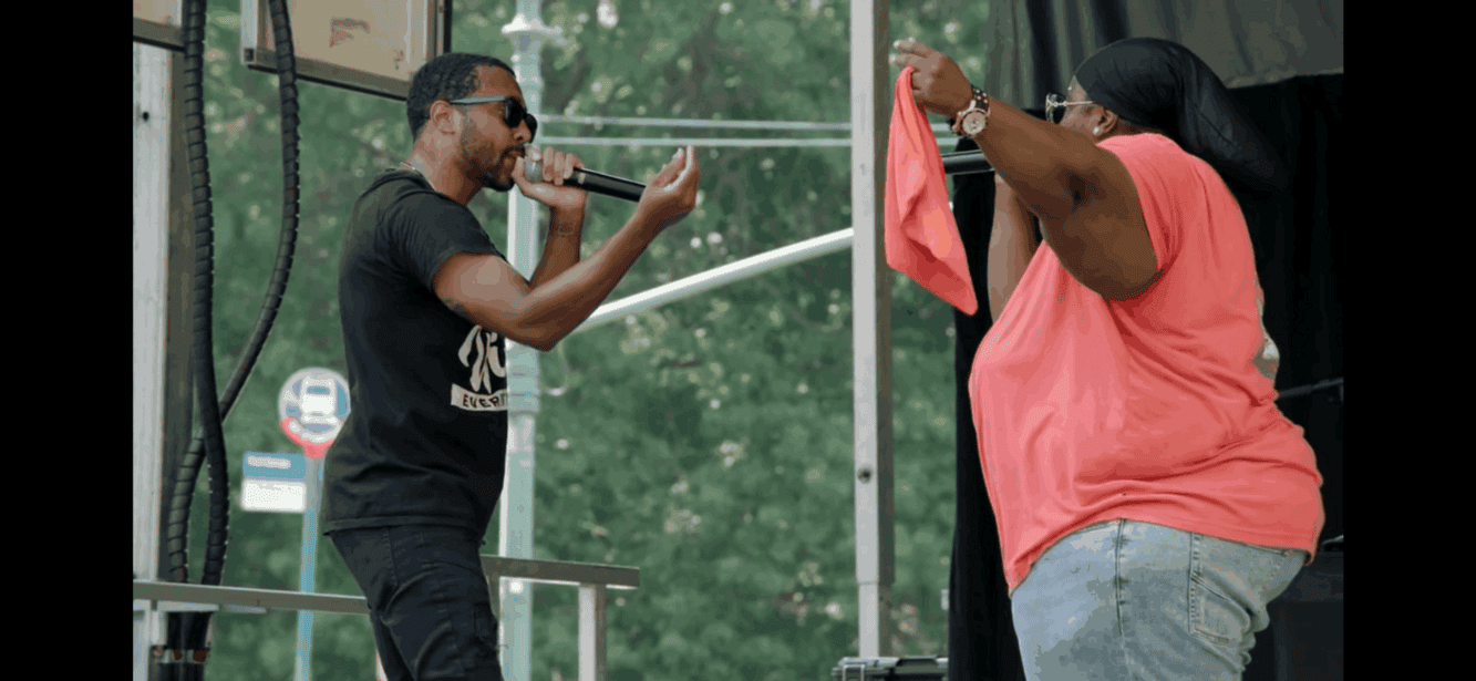 Man performing on stage with microphone and woman holding cloth outdoors, live music event.