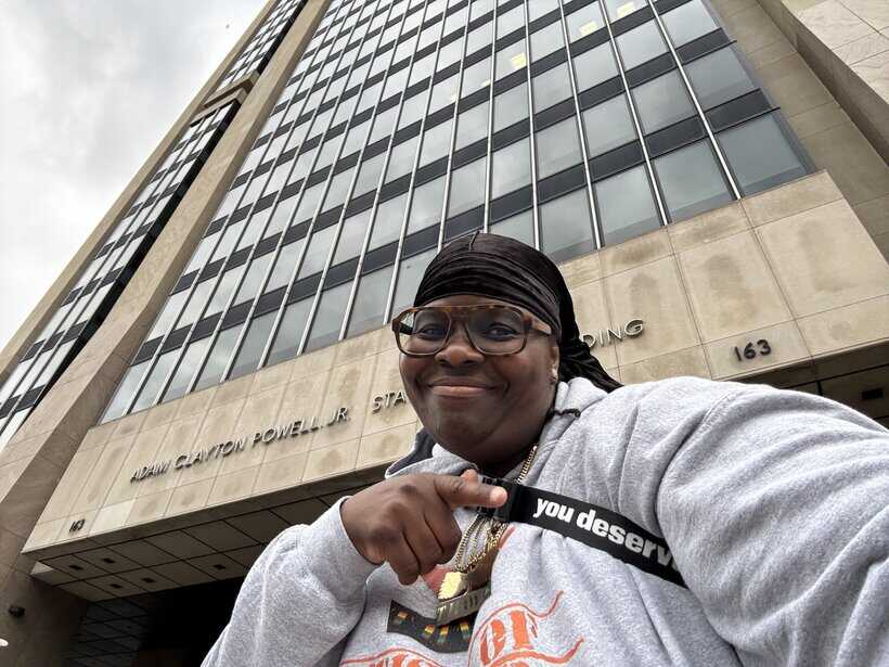Smiling person taking selfie in front of Adam Clayton Powell Jr. State Office Building in NYC.