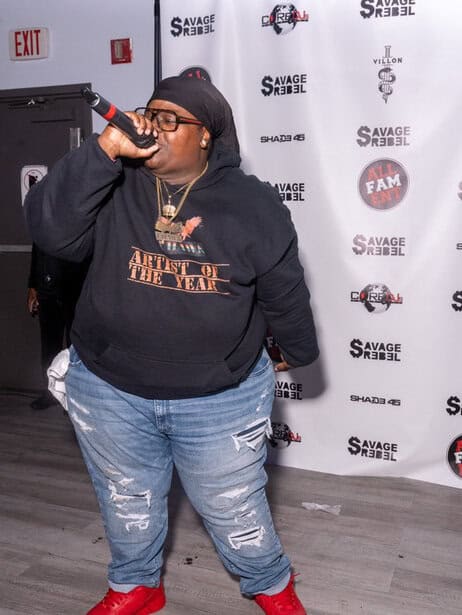 Unisex performer with microphone at music event, casual style, large hoodie and ripped jeans, urban atmosphere.