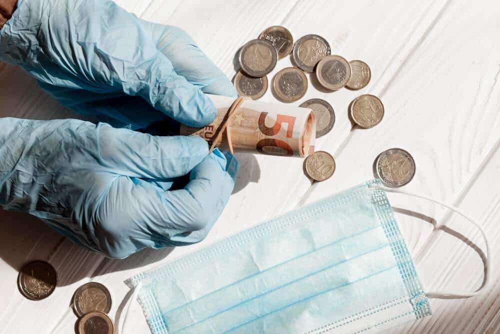 Hand hält Euro-Banknote mit Münzen, medizinischer Maske und Handschuhen auf weißem Hintergrund.