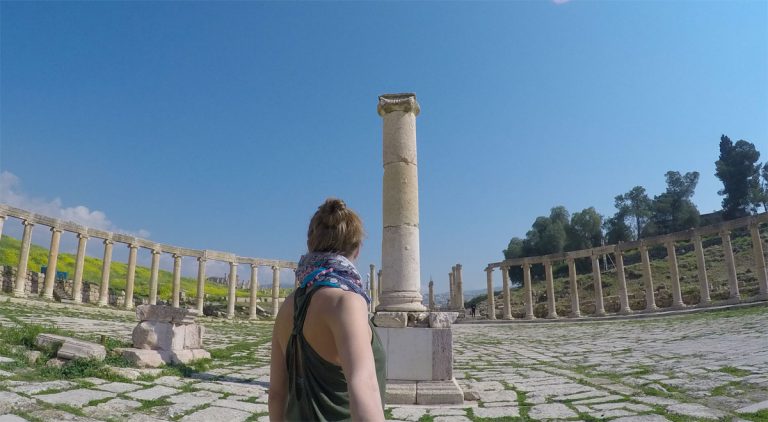 A woman taking a selfie while admiring the beautiful ruins of the Forum, featuring a tall column at the center and multiple columns lined in a circular row, with a blue sky in the background