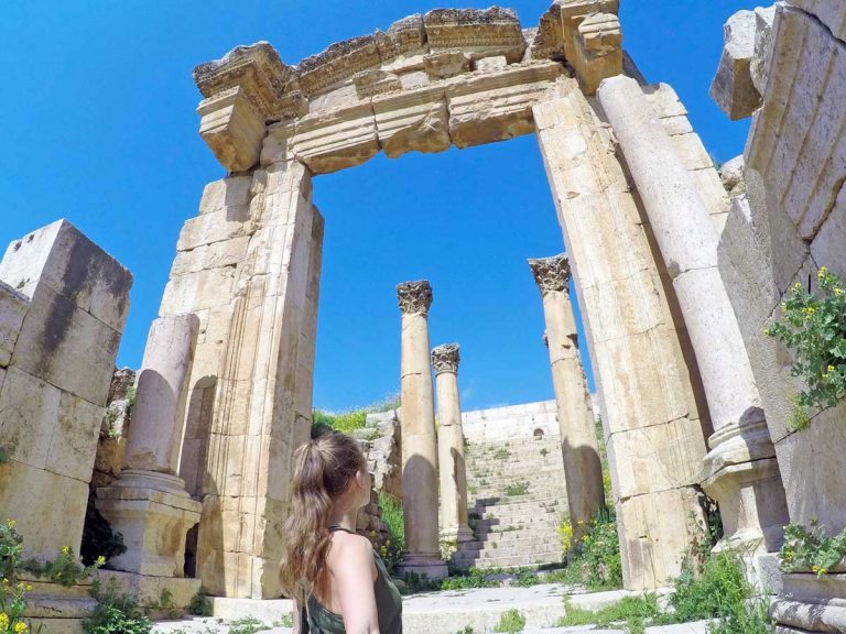 A woman taking a selfie while admiring the Ruins of Jerash in Jordan, featuring an old arch and multiple tall columns on a sunny day