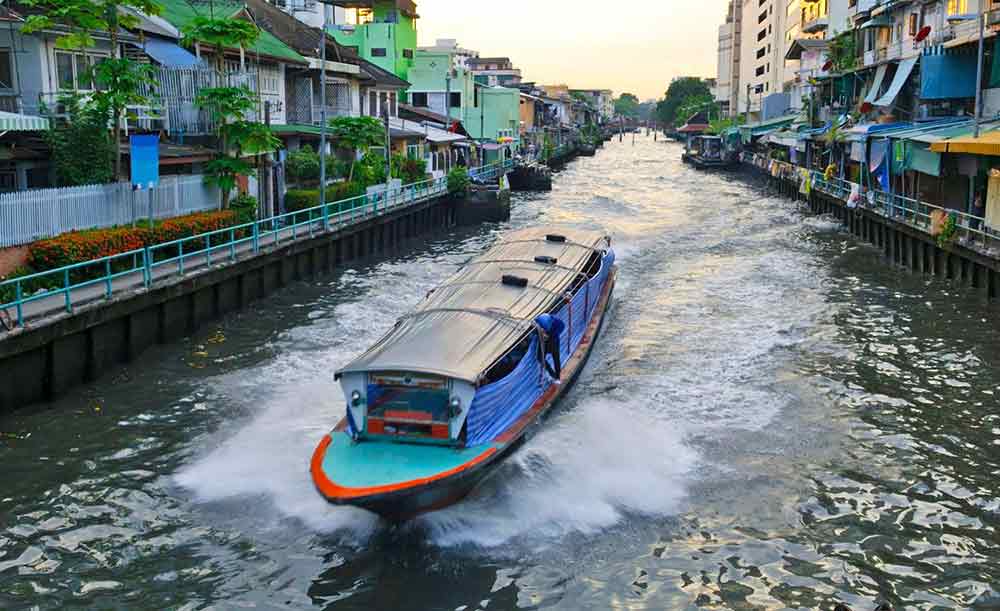 A water taxi in Bangkok going along a canal with houses and buildings at the sides. 
