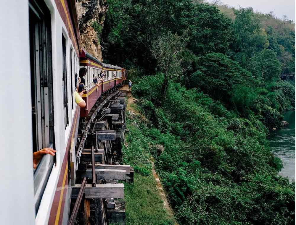 The old train going along the Death Railway bridge with trees and a river to one side. 