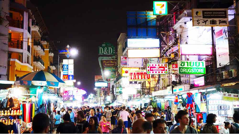 Khao SanRoad in Bangkok filled with neon lights and vendors selling food and other products with lots of people shopping. 