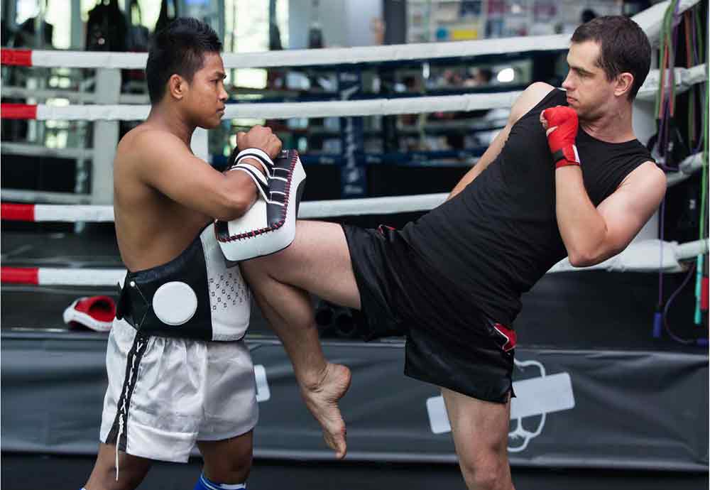 A Muay Thai lesson with a man in a black tshirt raising his knee up to a pad held by a teacher. 