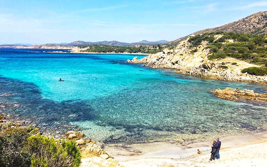 33 Best Hidden Coves and Secluded Beaches Around the World 23 A couple stands in awe, taking in the beauty of Cala Antoniareddu in Sardinia, Italy; the clear blue waters reveal the stones and sand beneath, creating a mesmerizing scene of natural wonder.
